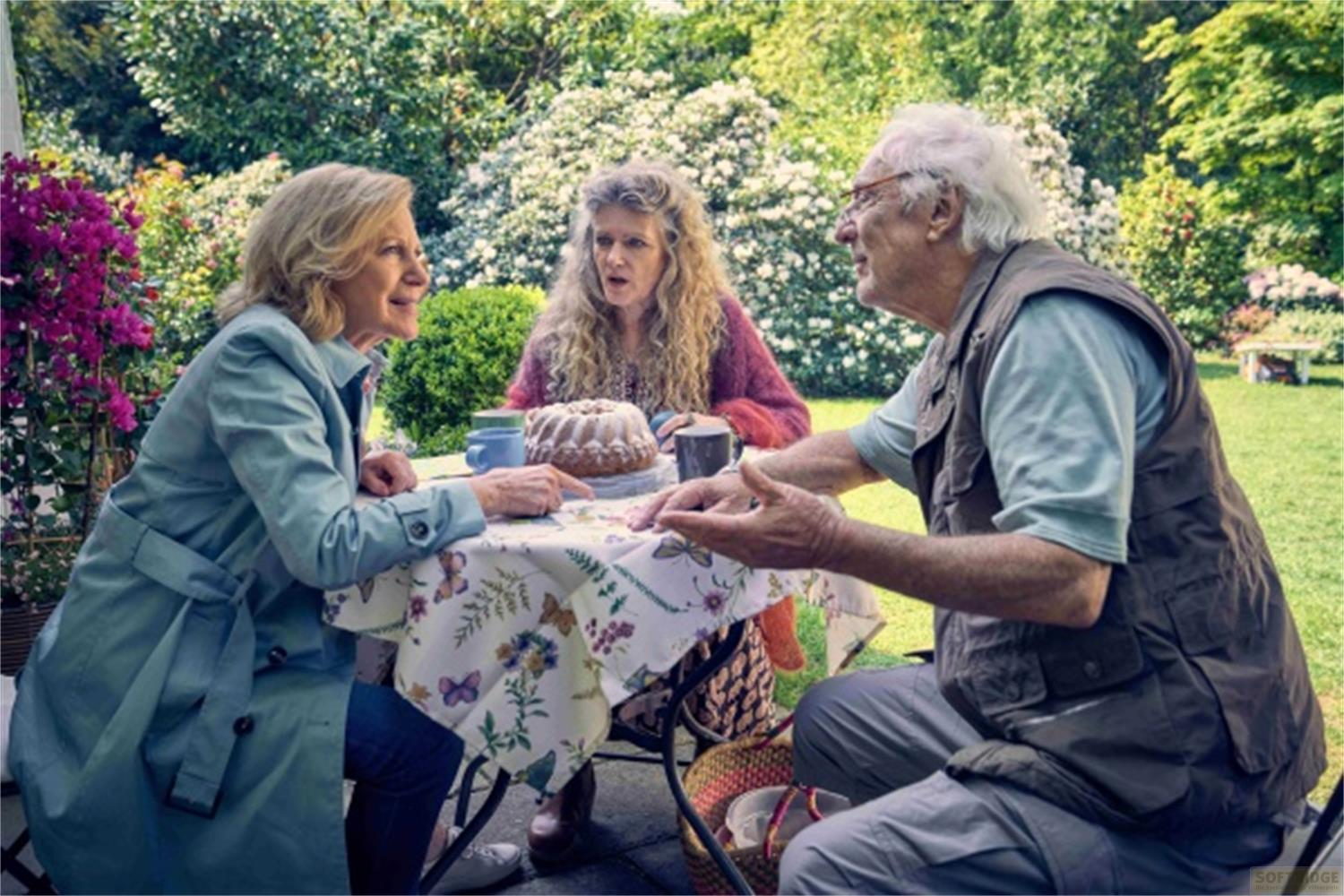 Tre persone sedute attorno a una torta in un giardino.
