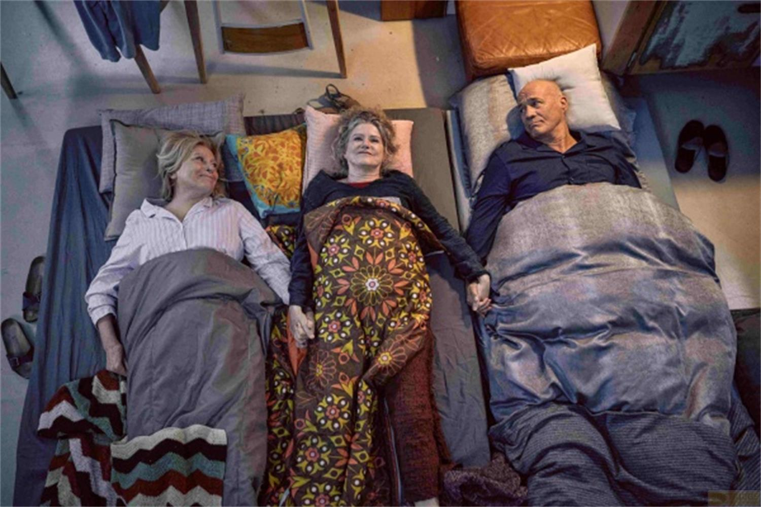 Tre persone giacciono fianco a fianco nel letto, tenendosi per mano.