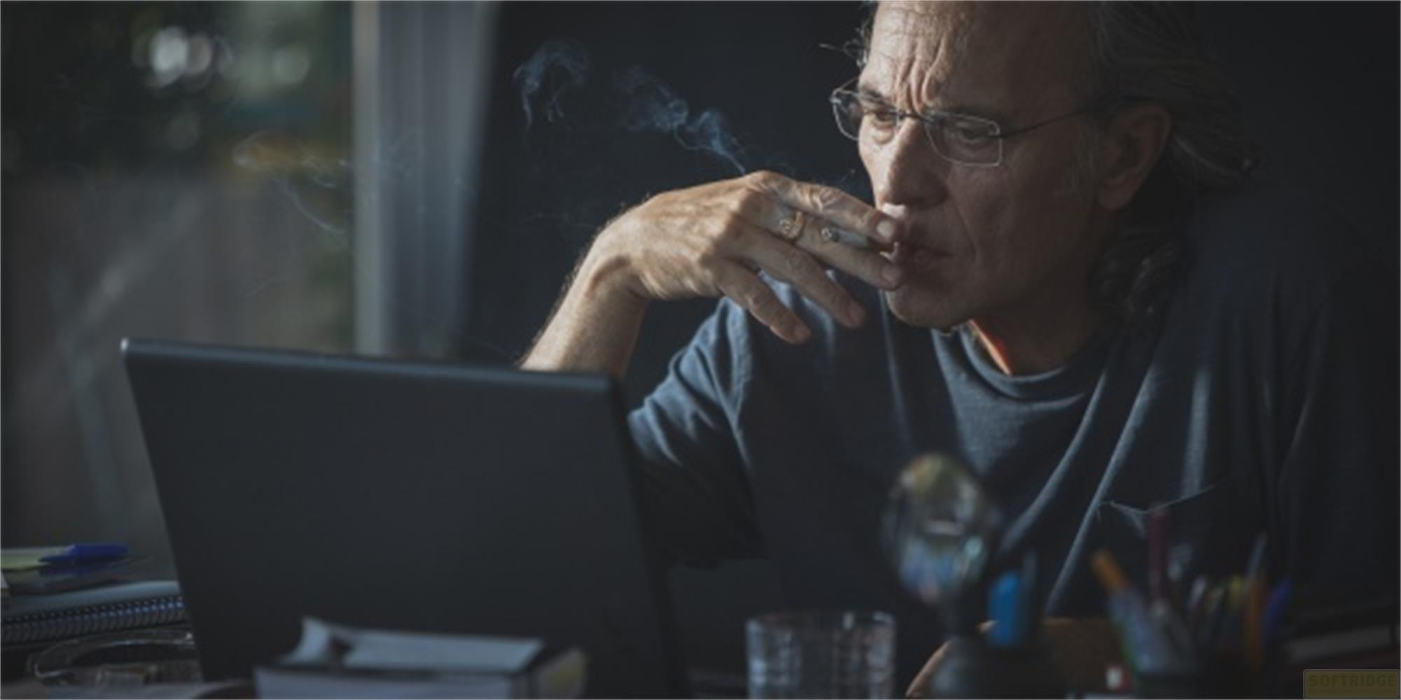 Un uomo con gli occhiali fuma guardando un laptop.