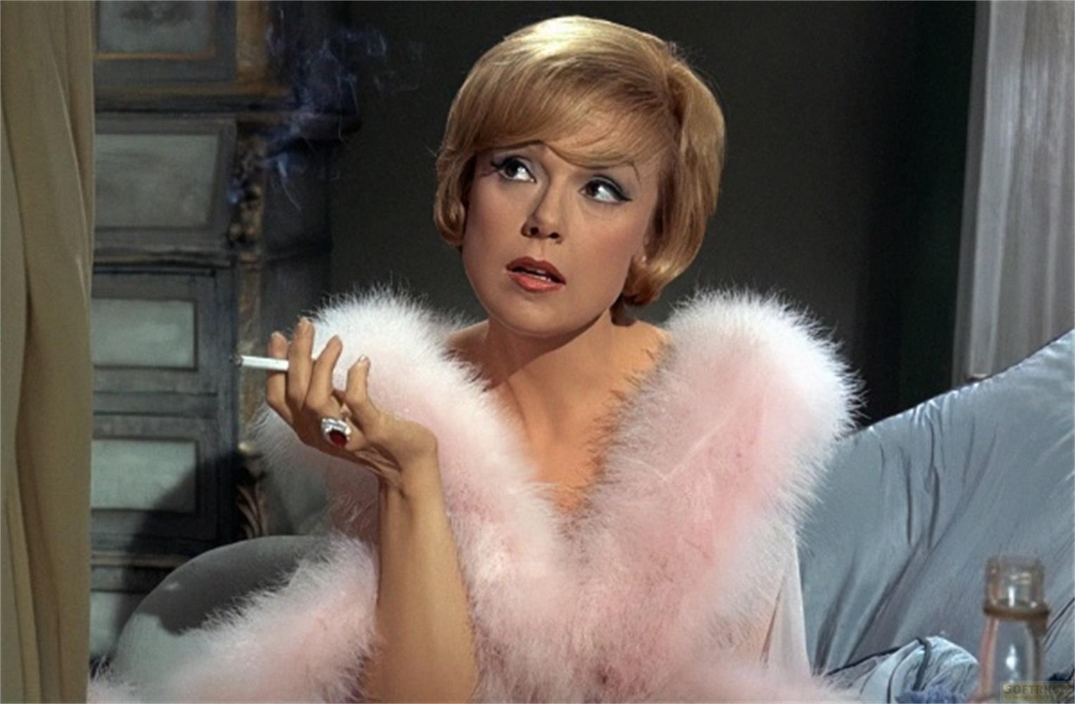 Donna bionda con capelli corti fuma sigaretta, indossa boa di piume rosa.