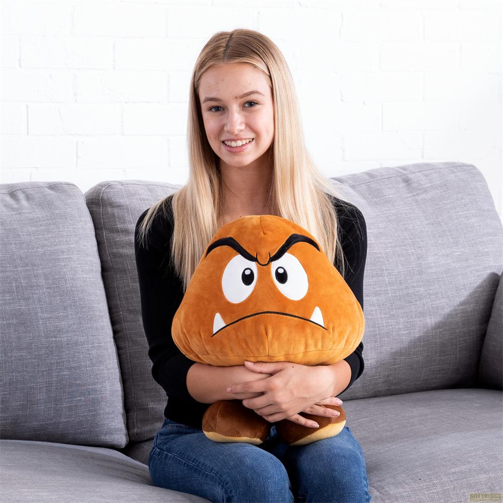 Eine junge Frau mit blonden Haaren sitzt auf einer grauen Couch und hält ein großes, braunes Goomba-Stofftier.