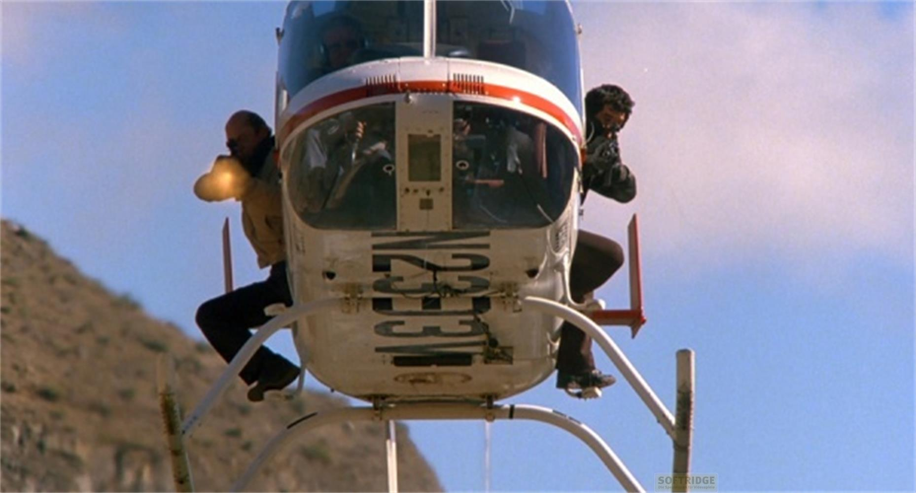 Deux hommes s'accrochent à un hélicoptère volant devant un fond montagneux.