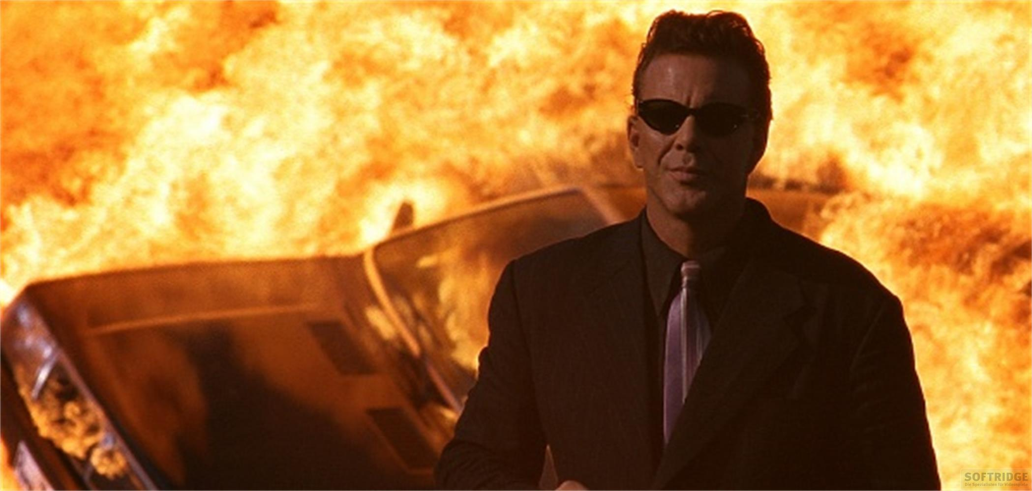 Un homme en lunettes de soleil et costume devant une voiture en feu.