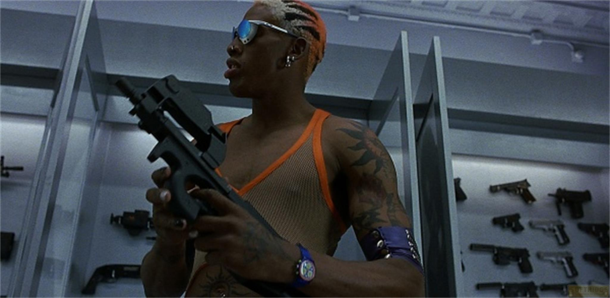 Un homme aux cheveux orange tient une arme, d'autres armes sont exposées au fond.