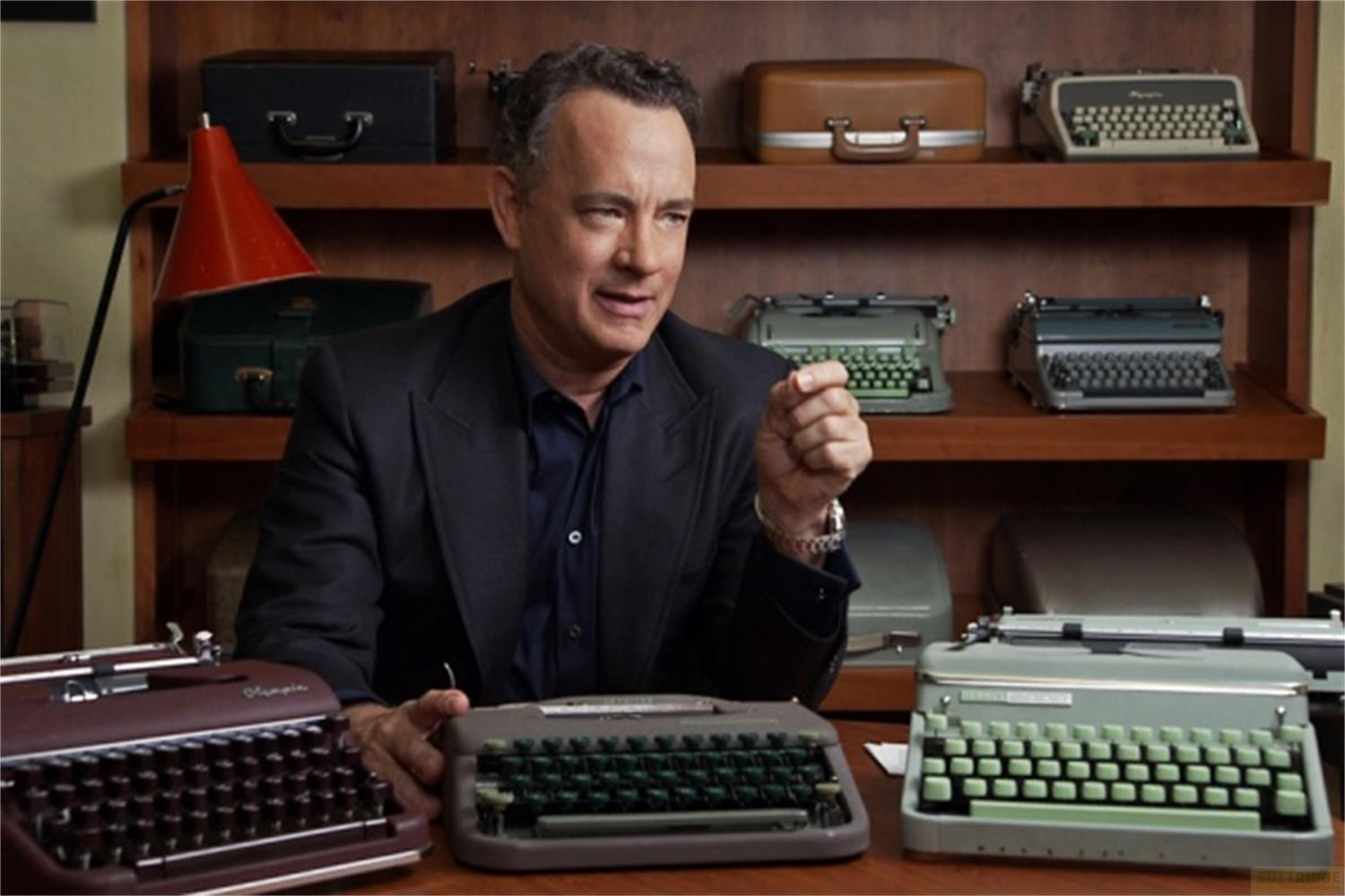Tom Hanks è seduto davanti a una collezione di macchine da scrivere. Indossa un abito.