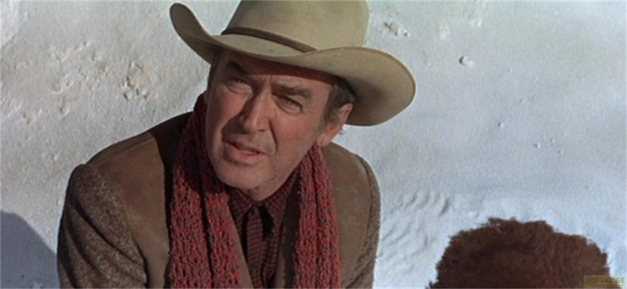 Un uomo con cappello da cowboy e sciarpa rossa parla davanti a un muro bianco.