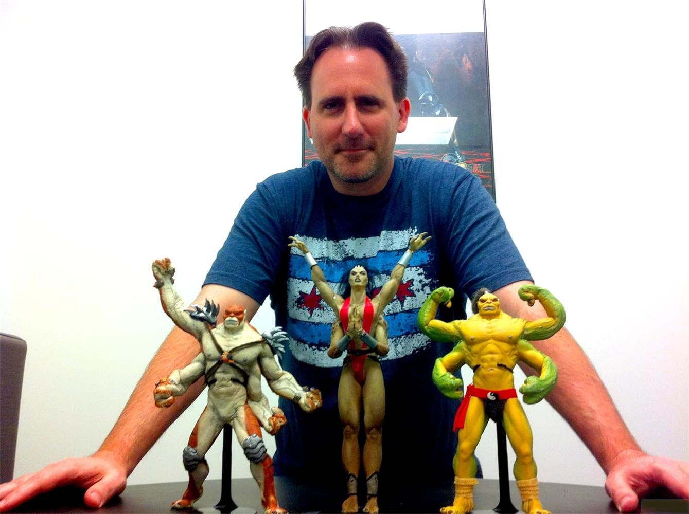 Mann mit drei Actionfiguren: links ein blasser Mann mit vier Armen, Mitte eine Frau, rechts ein gelber Mann mit vier Armen.