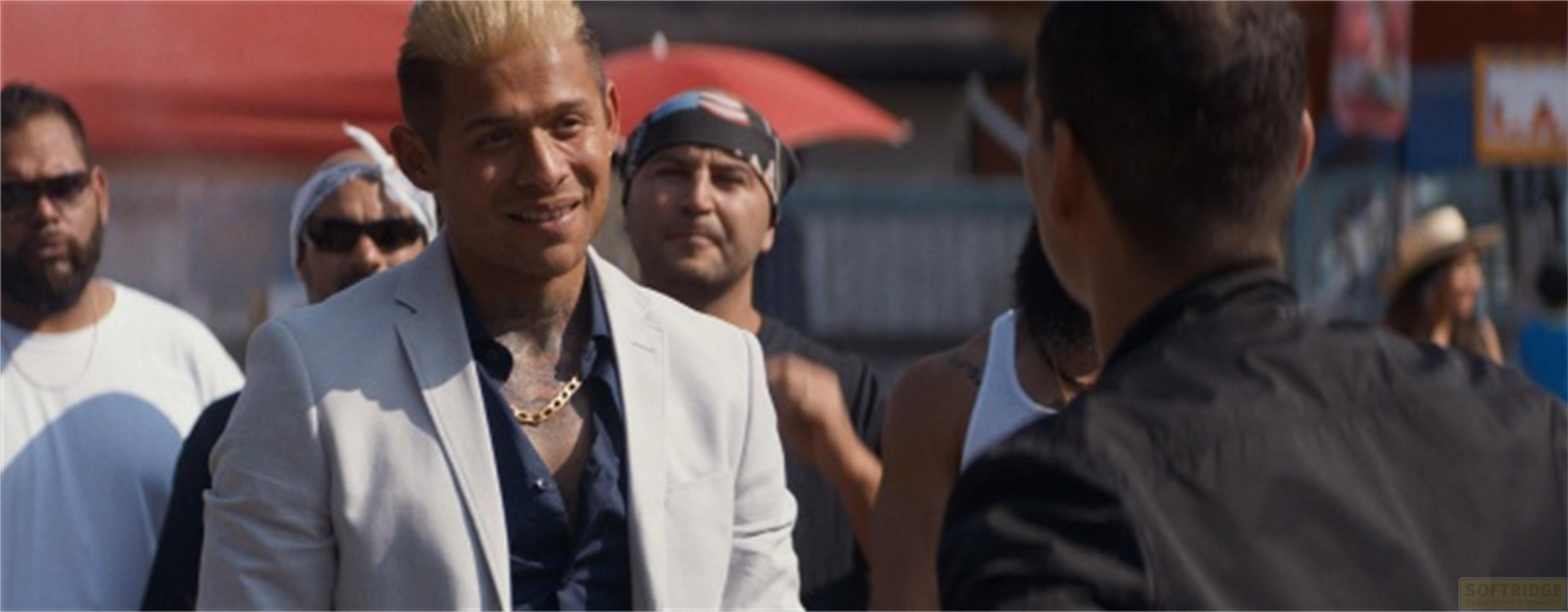 Ein Mann mit blond gefärbtem Haar trägt einen hellen Anzug und eine goldene Kette.