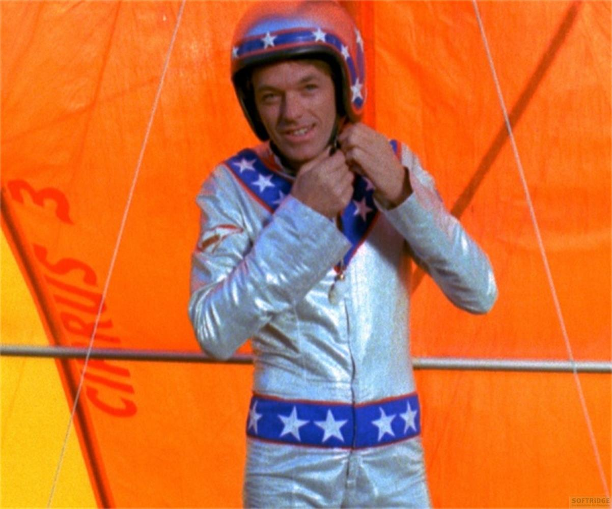 Un homme en costume argenté et casque à étoiles, devant une surface orange.