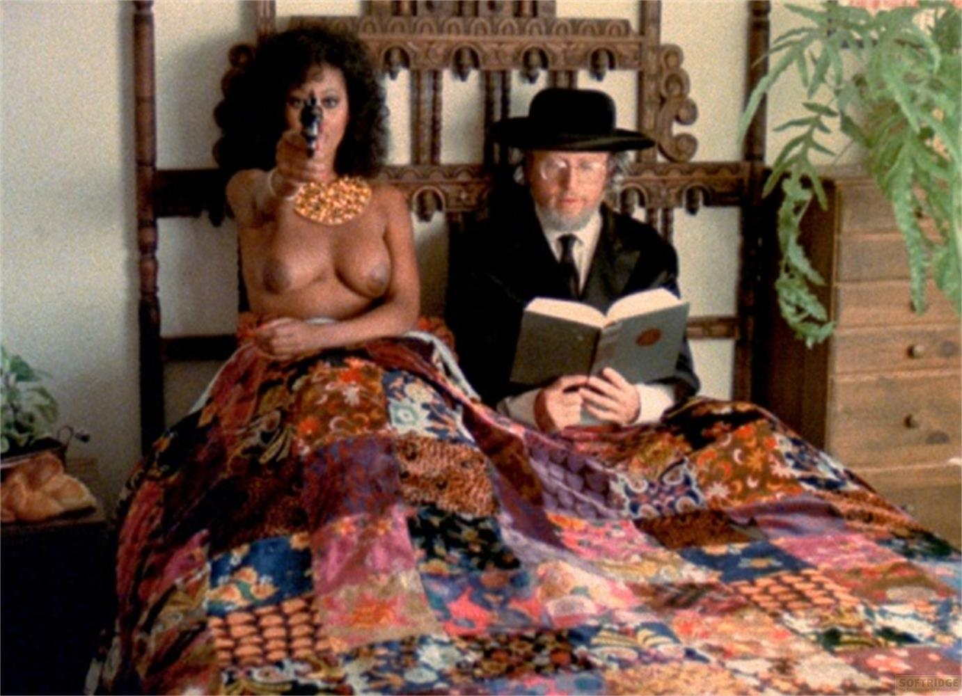 Une femme avec un pistolet et un homme avec un livre sont assis sur un lit avec une courtepointe.