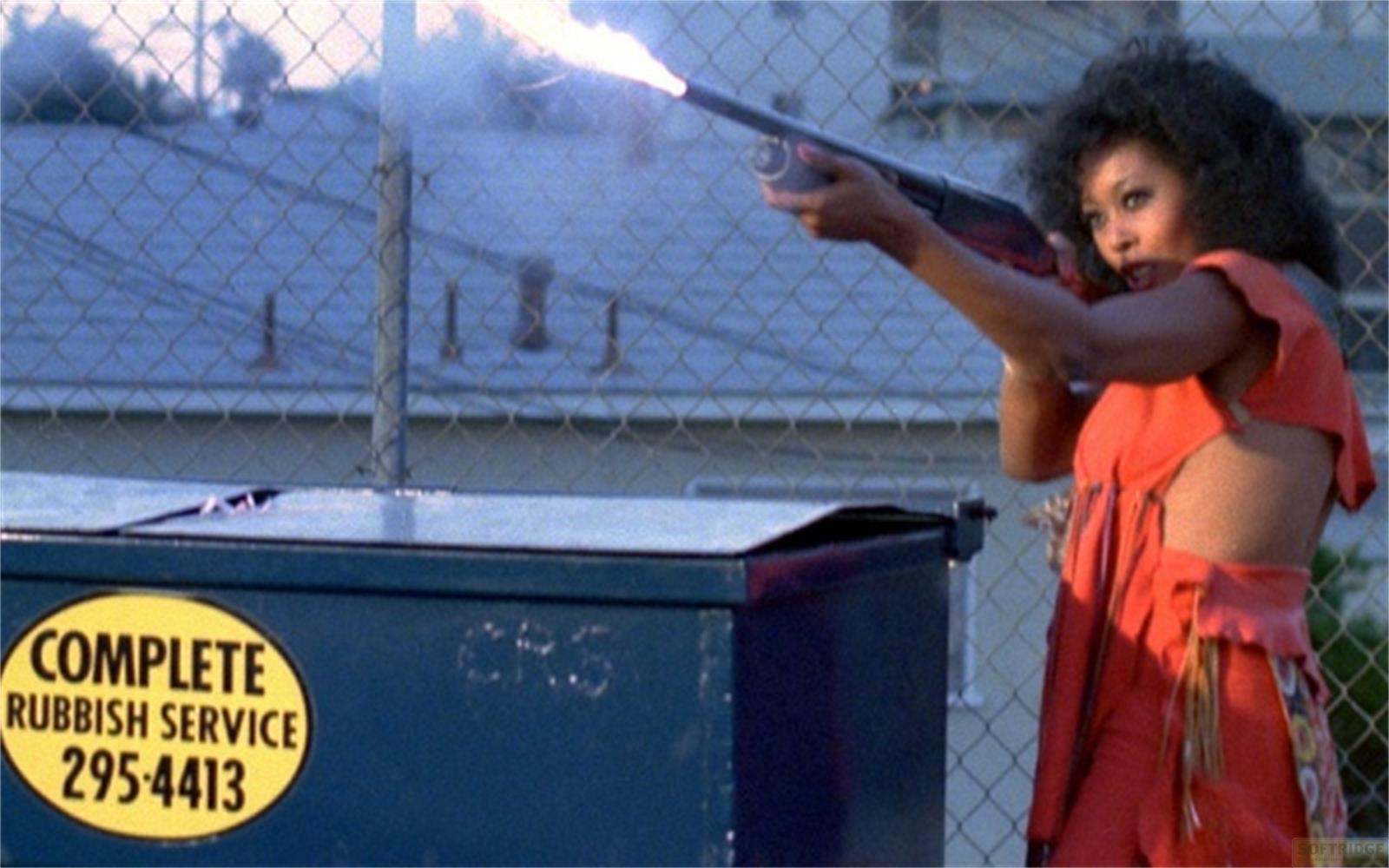 Une femme en tenue orange tire avec un fusil, de la fumée s'échappe.