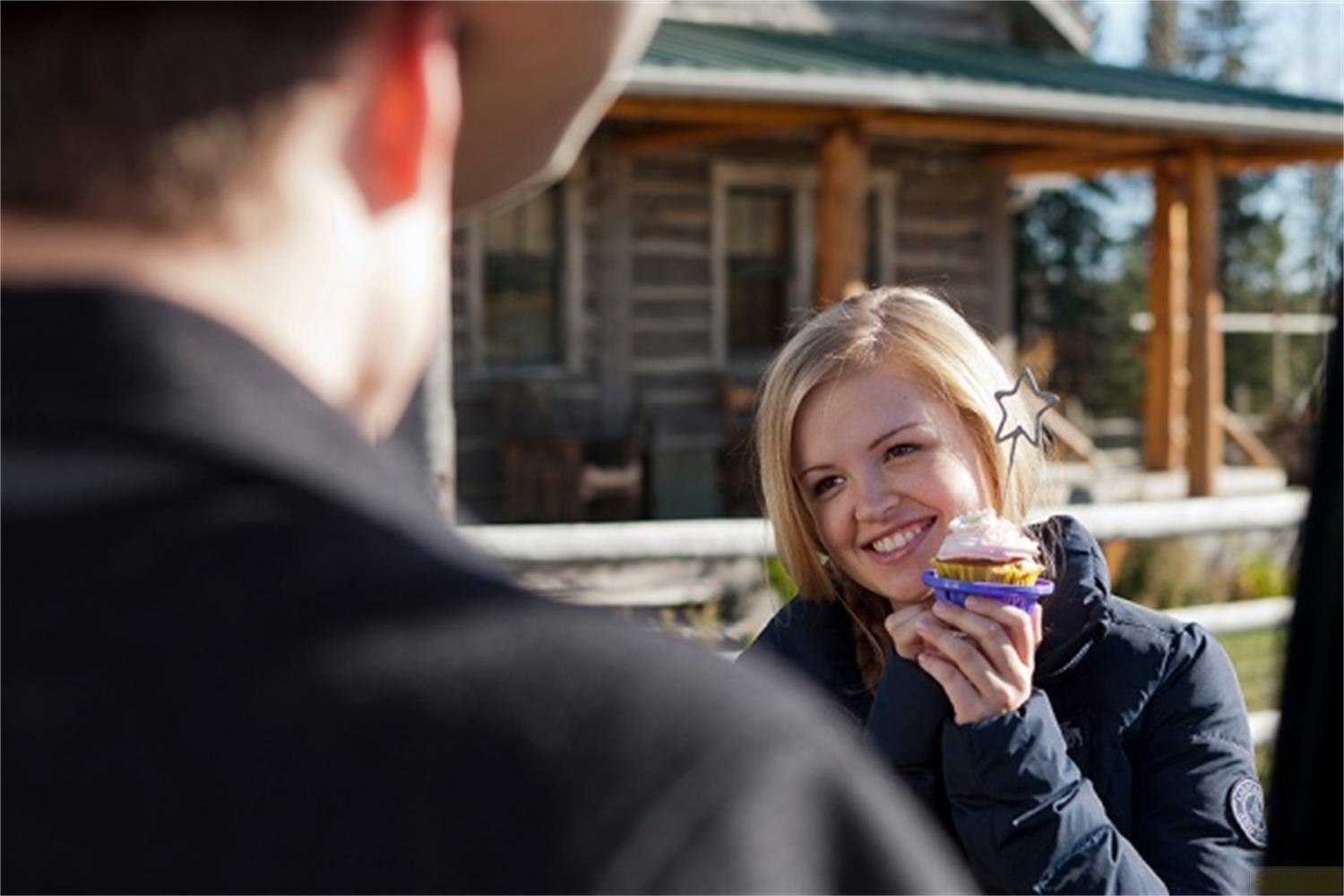 Une femme tient un petit gâteau devant une cabane en bois.