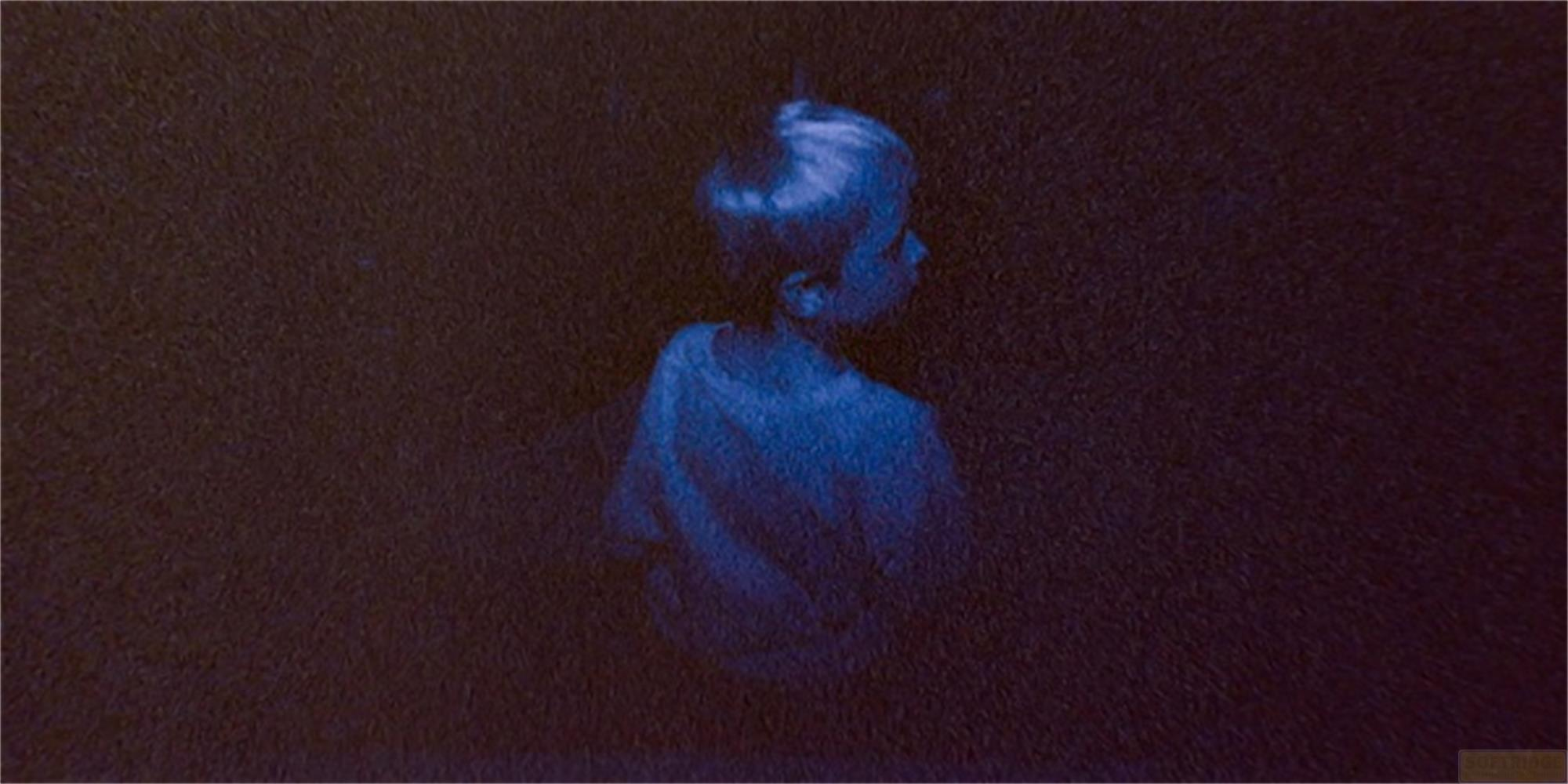Un ragazzo in camicia blu guarda a destra in un ambiente scuro.