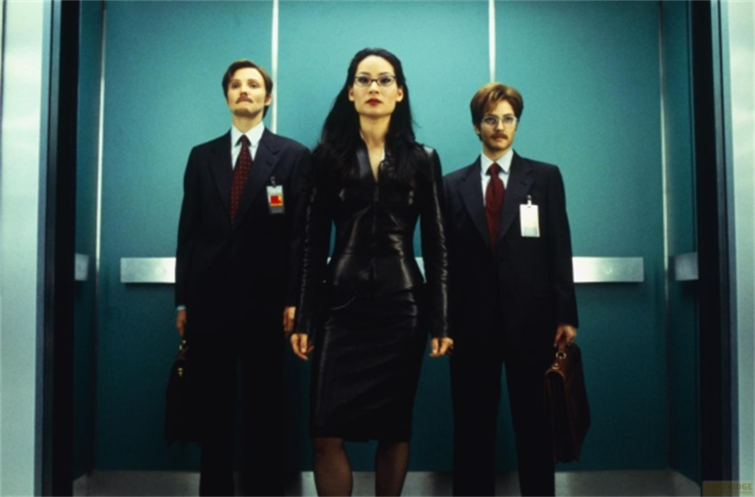 Trois personnes en costume devant un mur turquoise. Une femme en cuir.