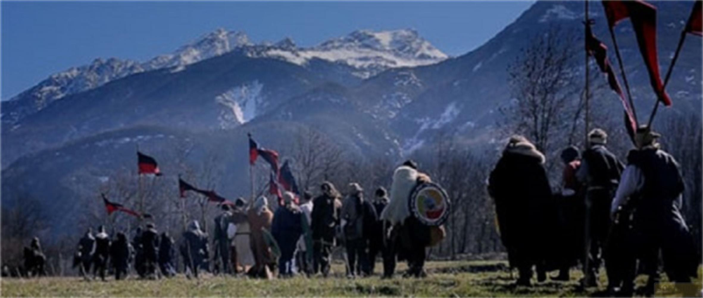 Un gruppo di persone in costume storico marcia con bandiere davanti a montagne innevate.