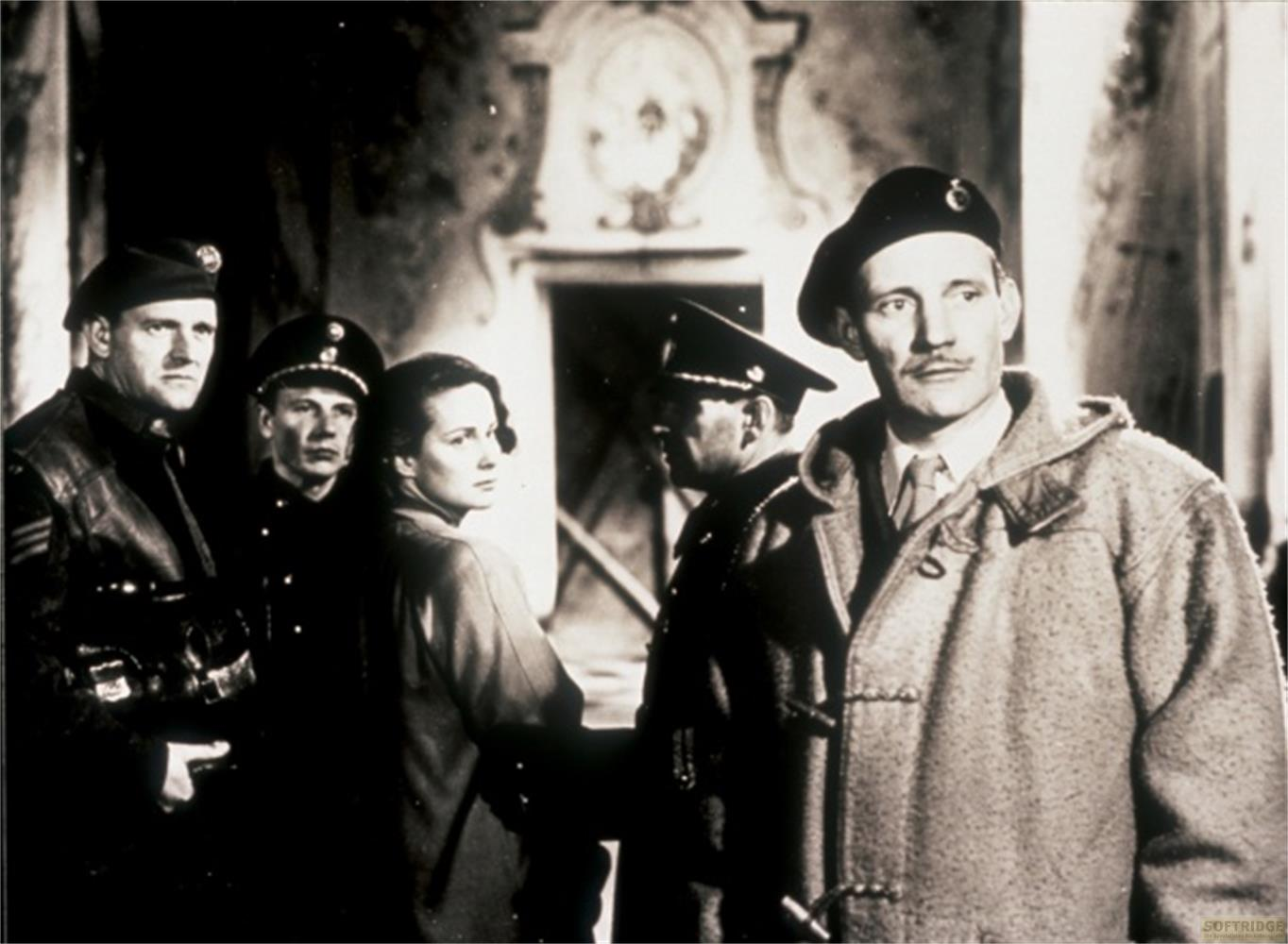 Männer in Uniformen und eine Frau stehen in einem Gebäude. Ein Mann trägt einen Dufflecoat.