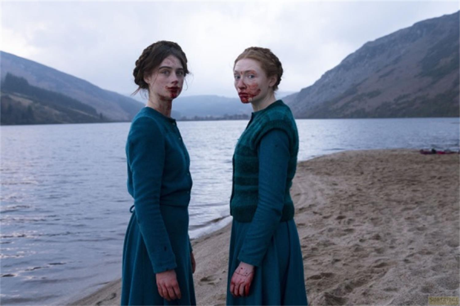 Due donne con il viso insanguinato vicino a un lago con montagne sullo sfondo.