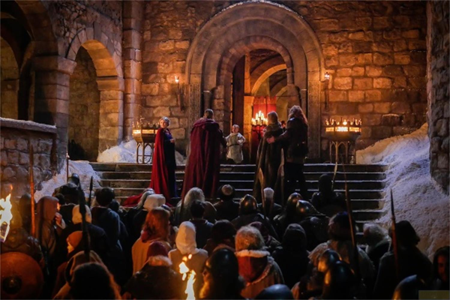Des gens en vêtements médiévaux se rassemblent devant des marches en pierre, éclairés par des torches et des bougies.