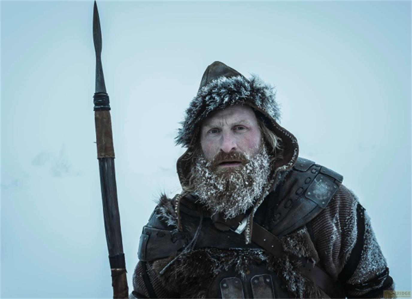 Un homme barbu et coiffé d'un bonnet en fourrure tient une lance dans un paysage glacial.