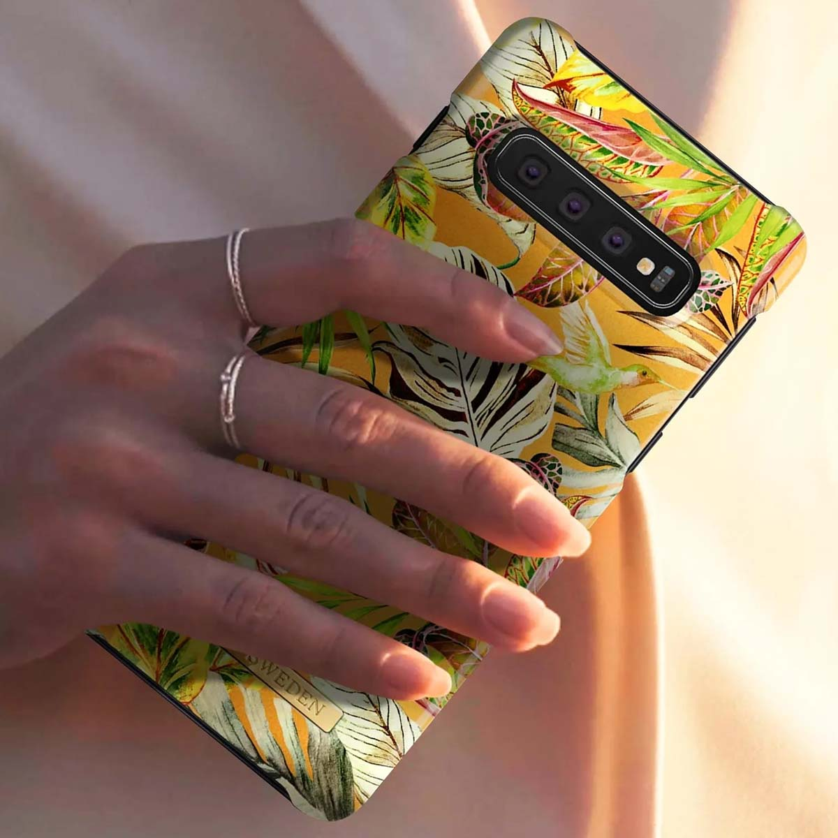 Una mano con anelli tiene un telefono con fantasia floreale tropicale e colibrì.