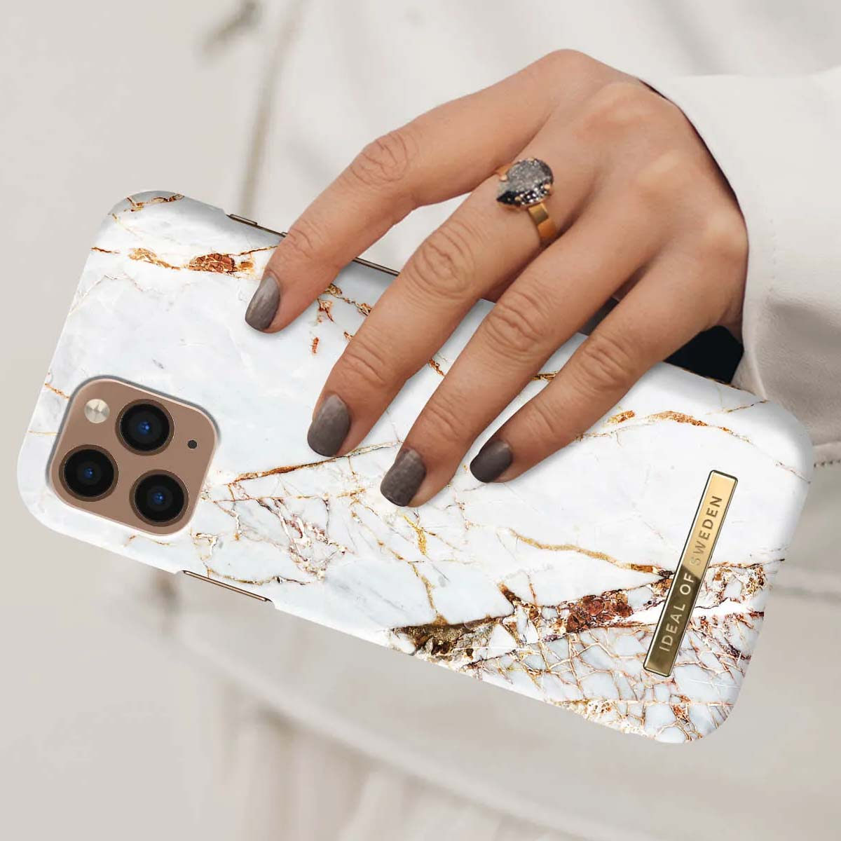 Una mano con esmalte gris sostiene una funda de mármol blanco con vetas doradas.
