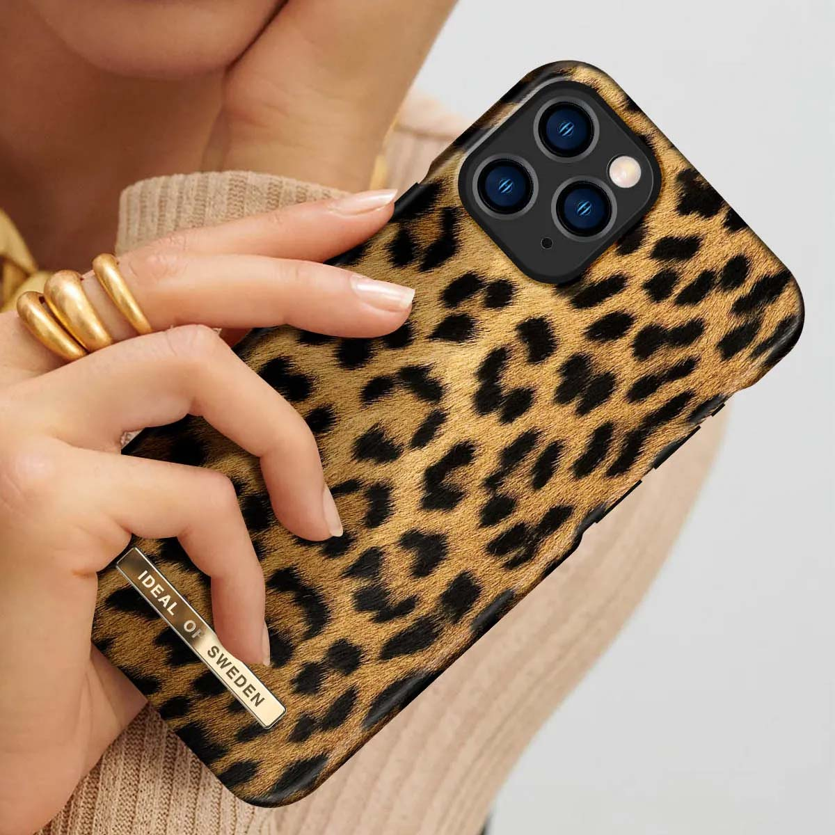 Una mano con anelli d'oro tiene un iPhone con stampa leopardata e logo IDEAL OF SWEDEN.
