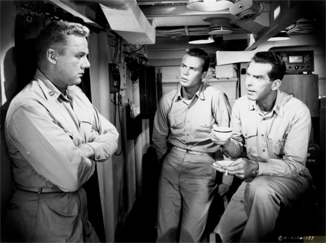 Trois hommes en uniforme conversent dans la cabine d'un navire. L'un est debout, bras croisés ; deux sont assis.