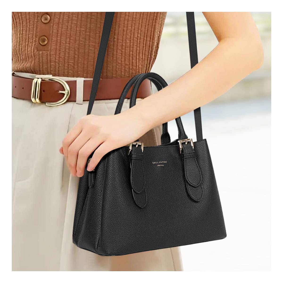 Schwarze Handtasche mit goldenen Schnallen, gehalten von einer Hand, neben braunem Gürtel.