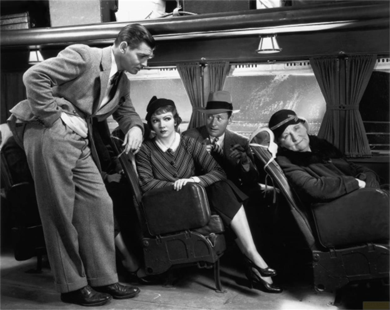 Un homme en costume parle à des passagers assis dans un train. Quatre personnes visibles.