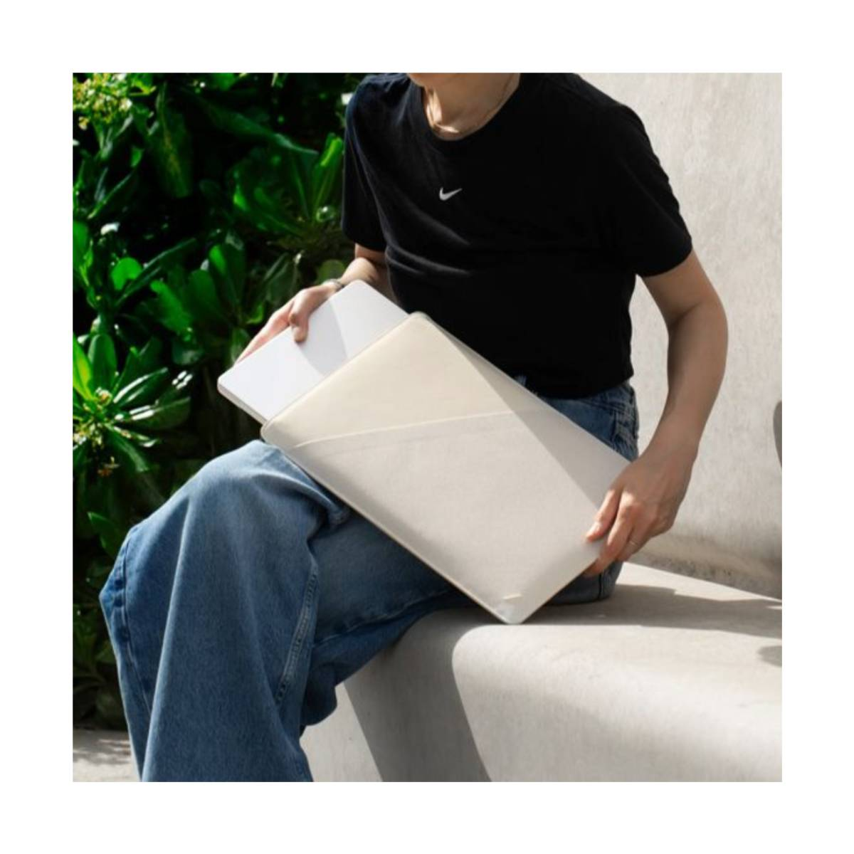 Una persona in jeans e maglietta nera tiene un laptop in una custodia beige chiaro.