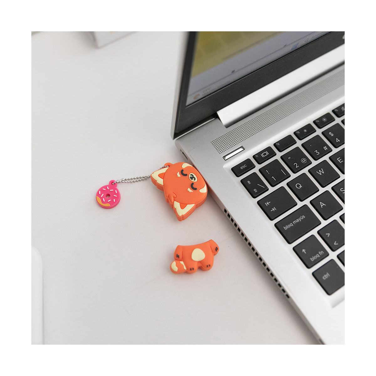 Ein orangefarbener USB-Stick in Fuchsform mit einem rosa Donut-Schlüsselanhänger neben einem Laptop.