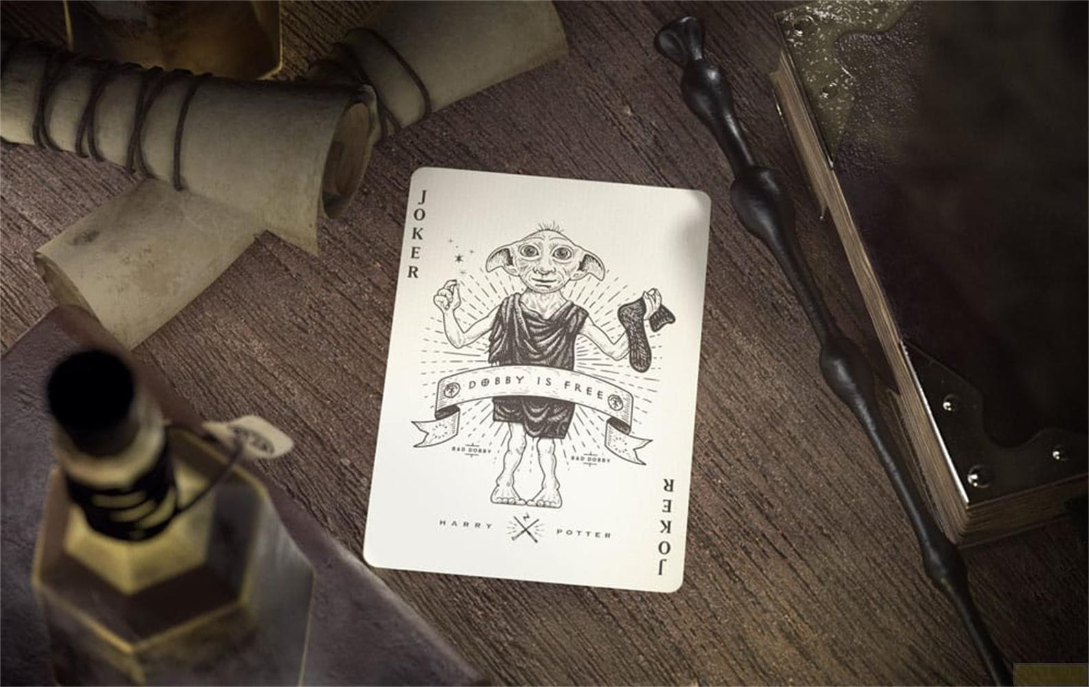 Carte Joker Dobby avec texte 'Dobby is free', baguette, bouteille et parchemins sur table en bois.