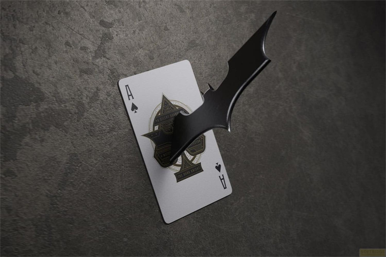 Un Batarang noir transperce une carte à jouer As de pique sur fond gris.