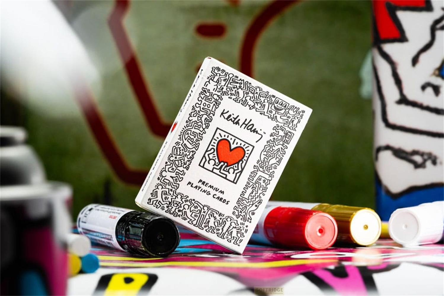 Boîte de jeu de cartes Keith Haring avec des marqueurs rouges, dorés et blancs sur une surface colorée.