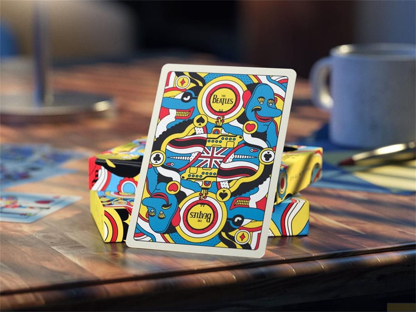 Beatles-Spielkarten mit buntem Design auf einem Holztisch, mit Tassen im Hintergrund.