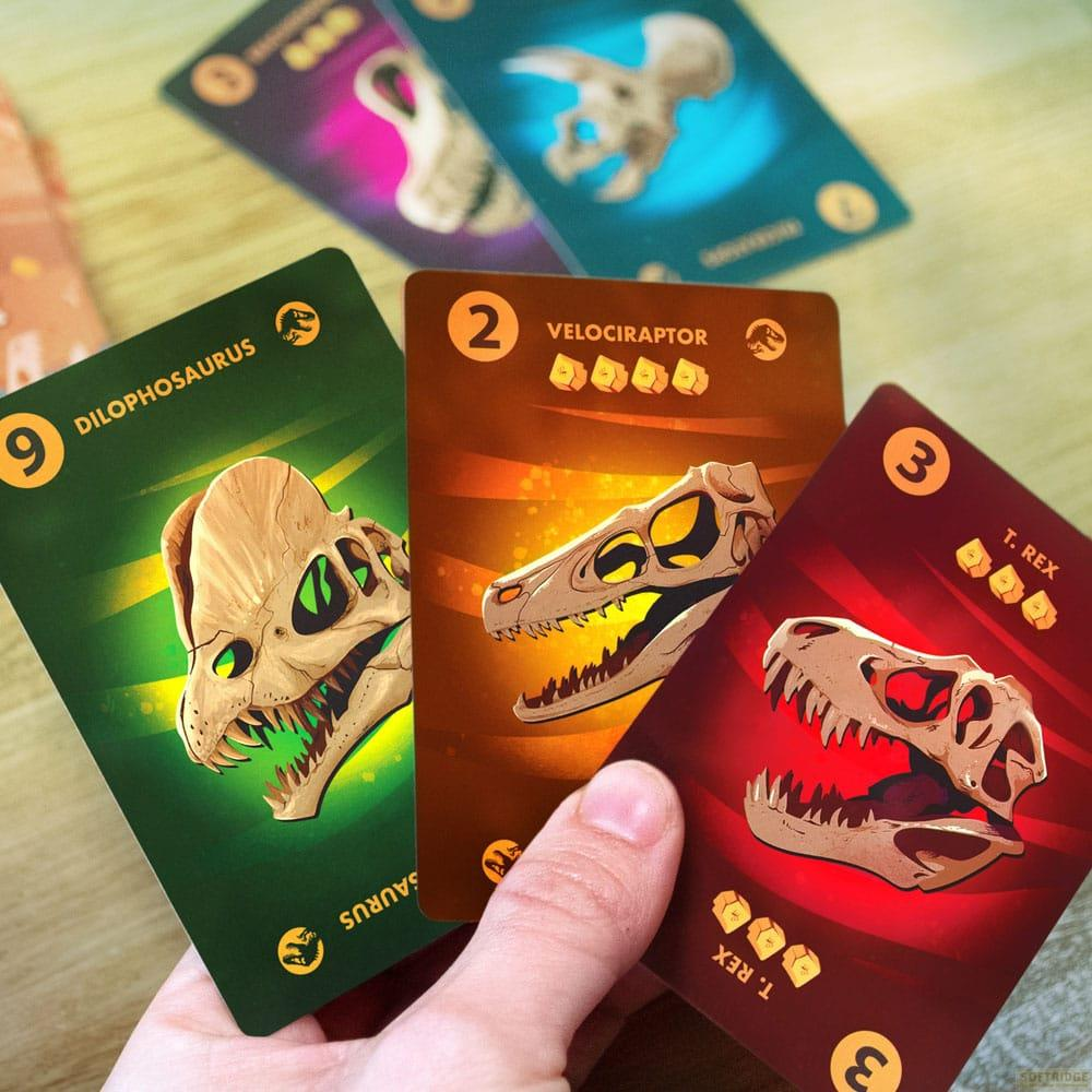 Trois cartes de dinosaures tenues par une main : Dilophosaurus, Vélociraptor, T. Rex.