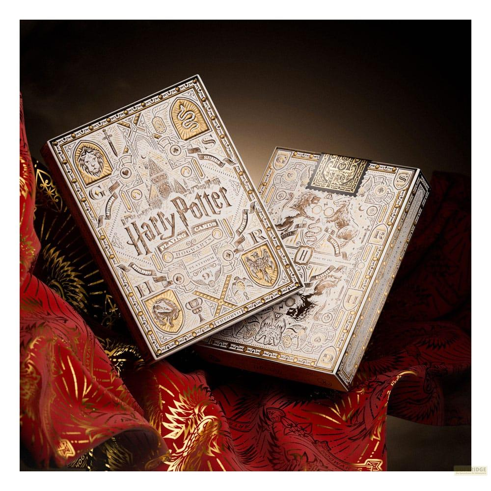 Deux boîtes de cartes à jouer Harry Potter ornées, blanches avec des motifs dorés, reposent sur du tissu rouge.