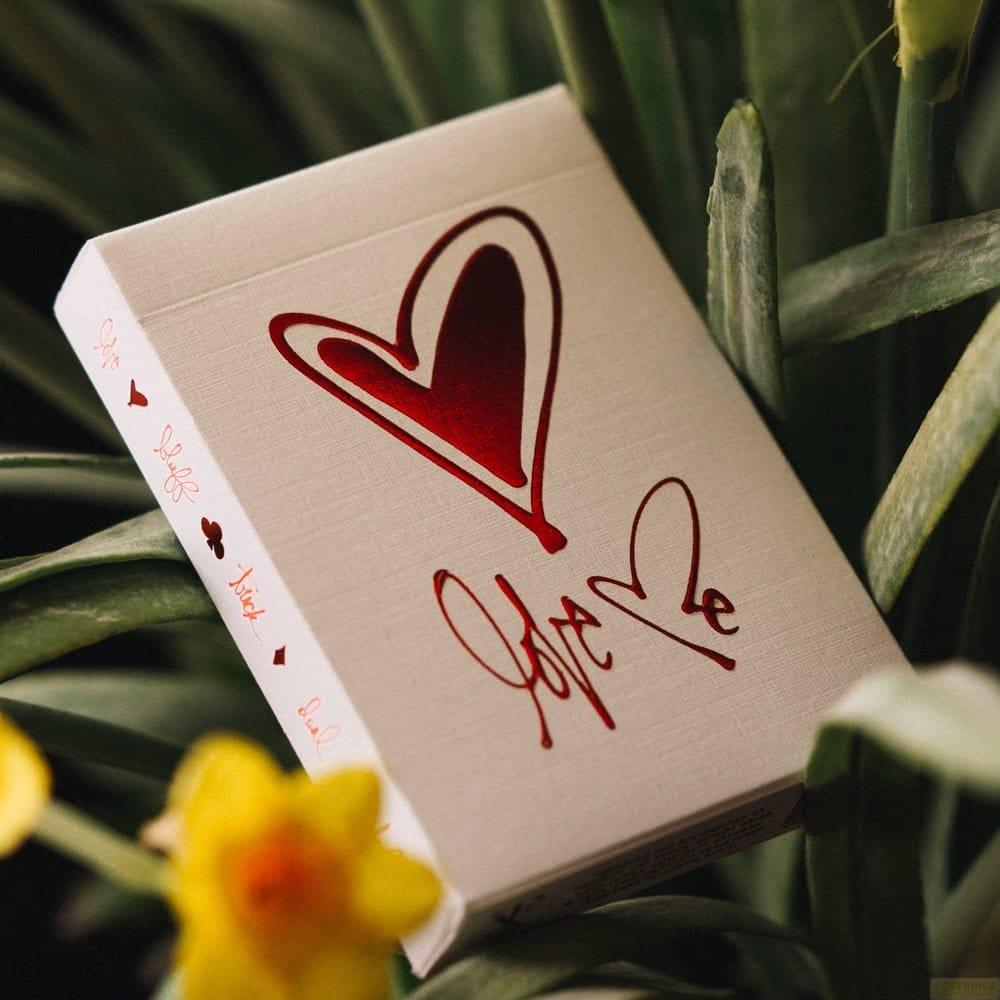 Un mazzo di carte color crema con cuore rosso e testo scritto si trova tra foglie verdi e fiori gialli.