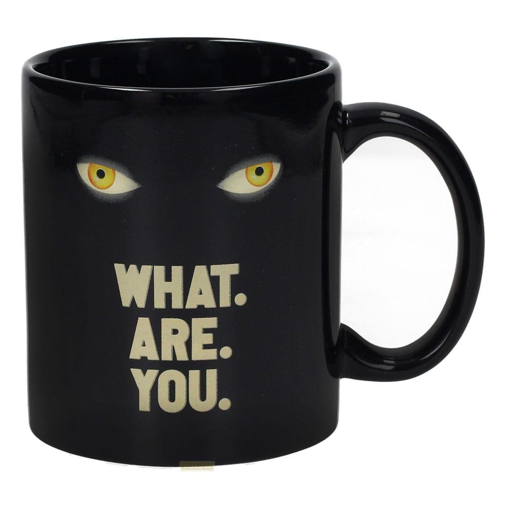 Una taza negra con ojos amarillos y el texto "WHAT. ARE. YOU.". Fondo blanco.