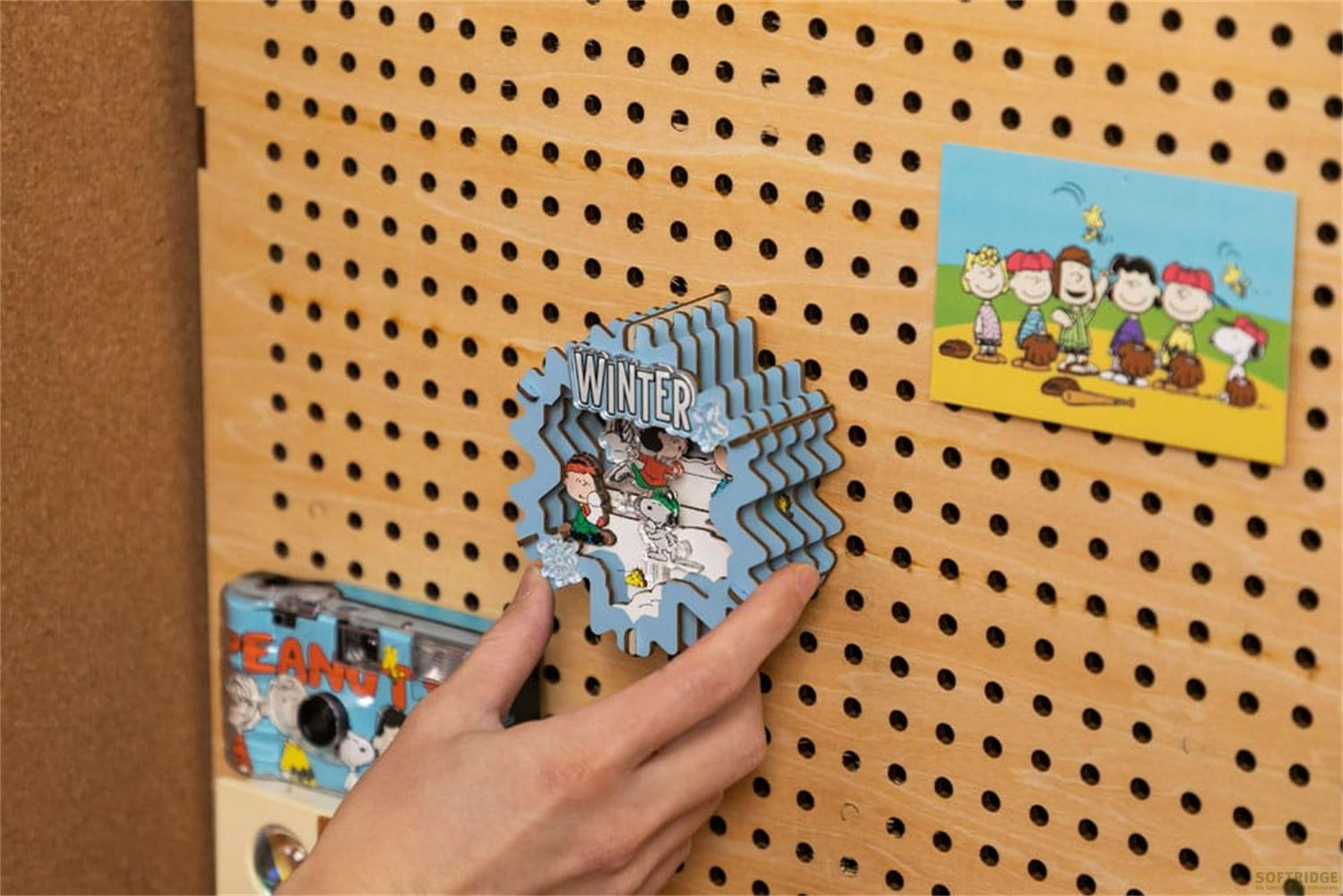 Une main tient un diorama bleu flocon de neige d'hiver avec des personnages de Snoopy sur un tableau à chevilles en bois.