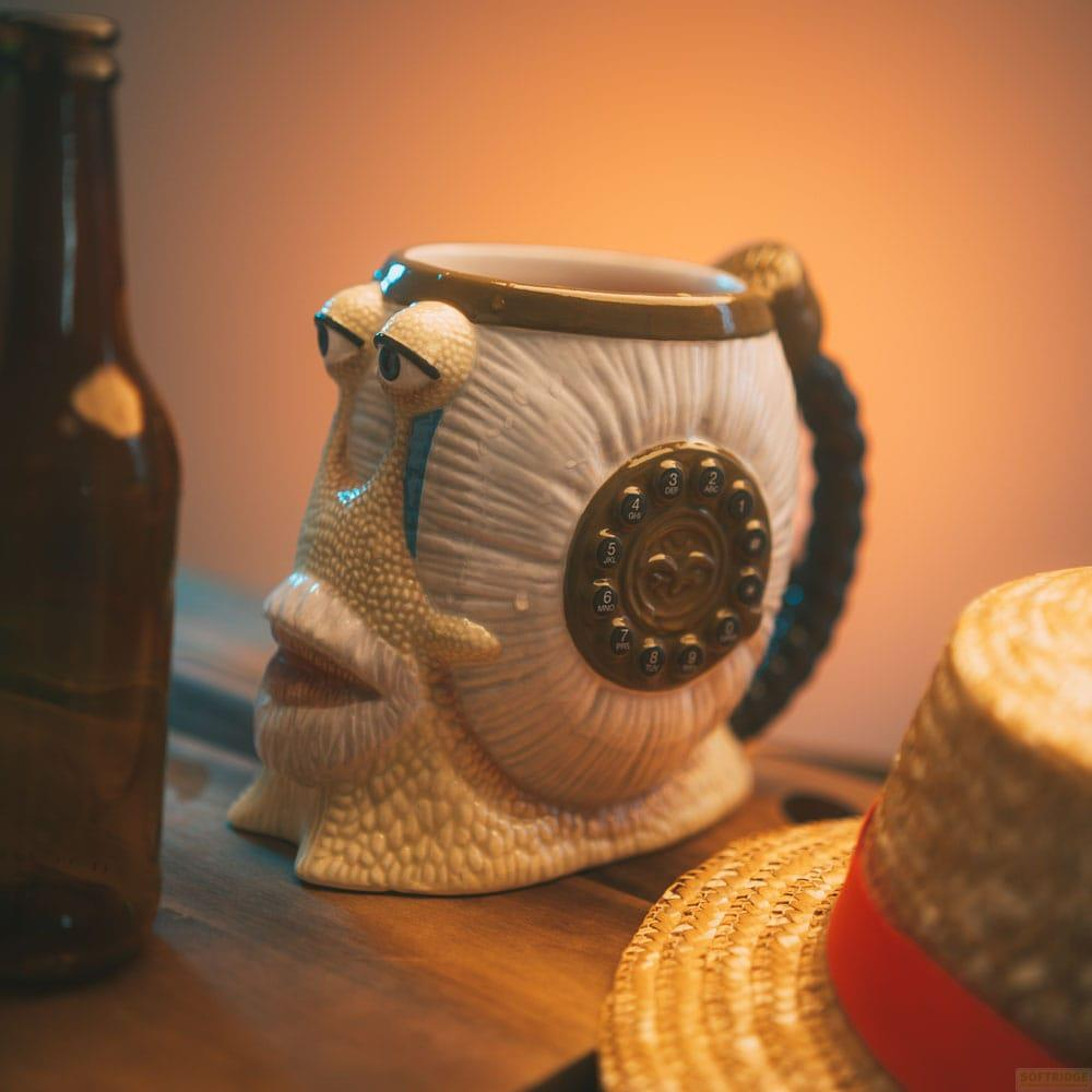 Keramiktasse in Schneckenform mit Telefonwählscheibe, daneben eine braune Flasche und ein Hut.