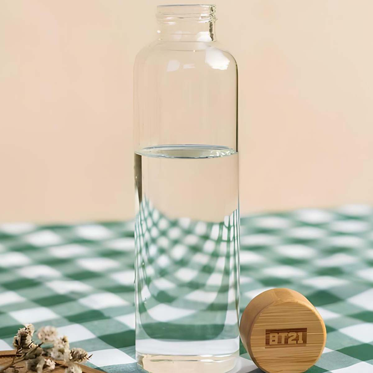 Una bottiglia di vetro con acqua è su una tovaglia a quadri verdi e bianchi, vicino a un tappo di legno con logo BT21.