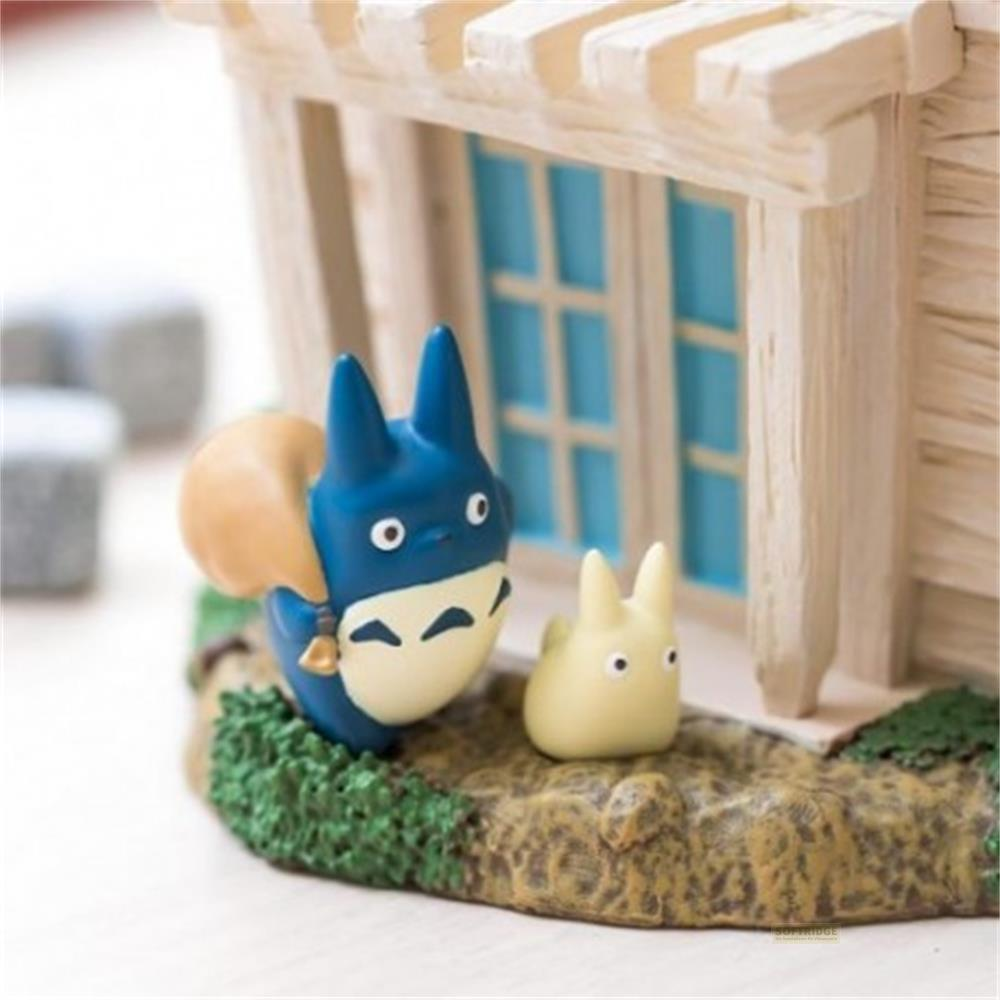Deux figurines Totoro devant une maison, une bleue avec un sac, une jaune à côté.