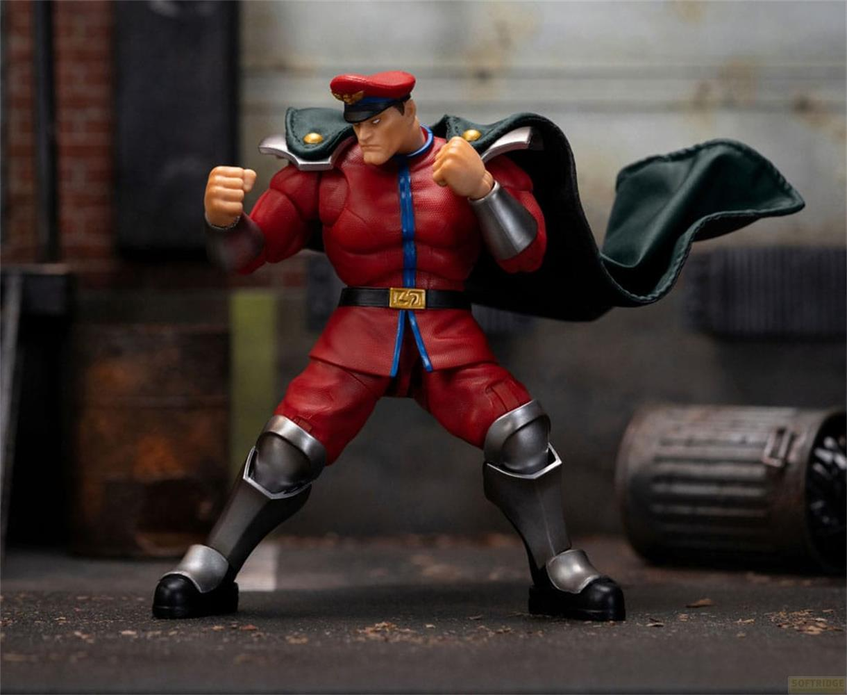 Eine M. Bison Actionfigur in roter Uniform, grüner Umhang, Fäuste geballt, steht auf einer staubigen Straße.