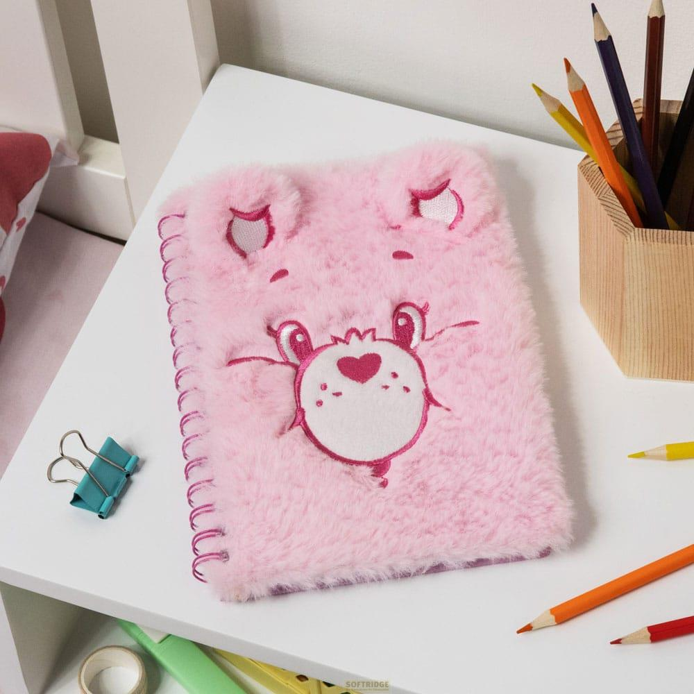 Ein flauschiges rosa Notizbuch mit einem Bären-Gesicht liegt auf einem weißen Tisch. Stifte sind daneben.
