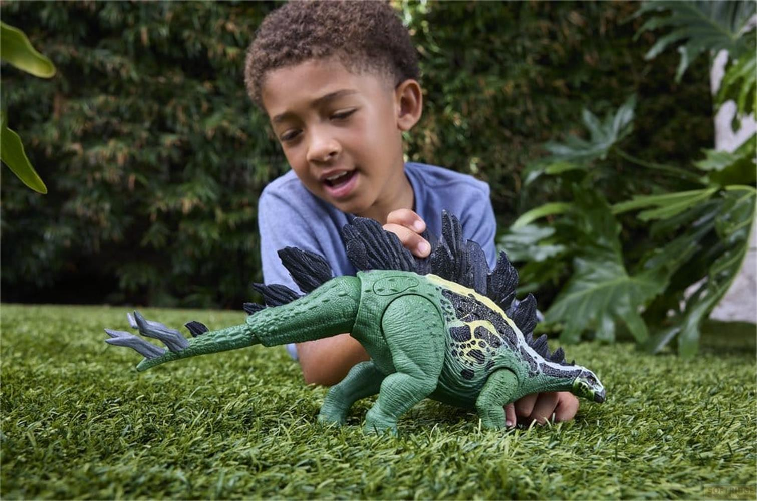 Ein Junge spielt mit einer grünen Dinosaurier-Figur auf einer Rasenfläche im Freien.
