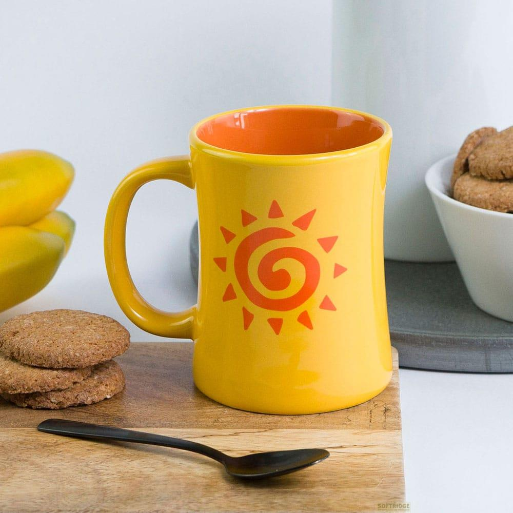 Taza amarilla con diseño de sol, junto a galletas y cuchara.