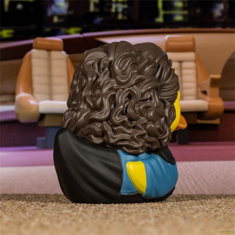 Eine Gummiente mit braunen Locken und blau-gelber Kleidung sitzt auf einem Teppich.