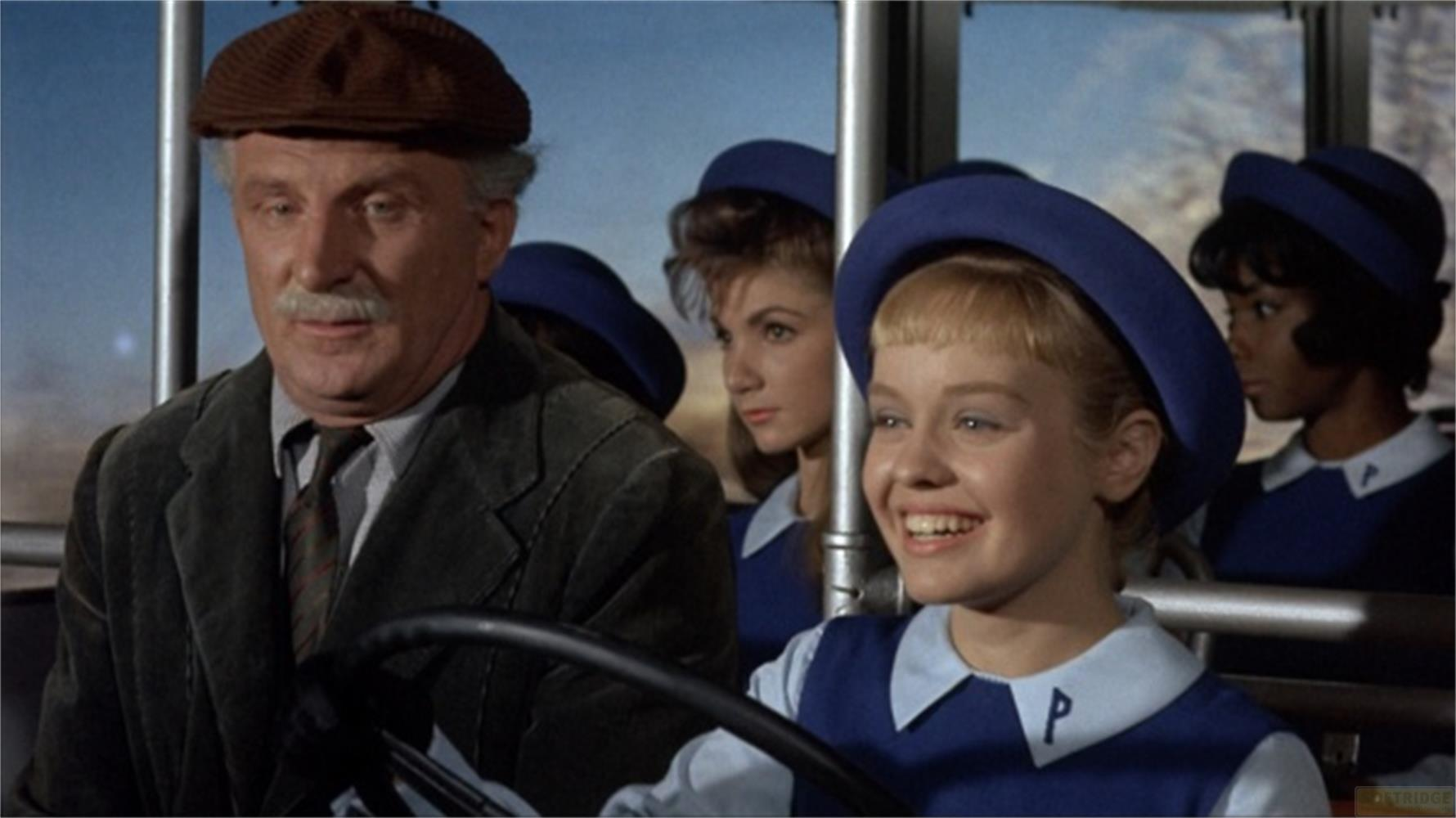 Un uomo con cappello marrone guida un autobus con ragazze in uniforme blu.