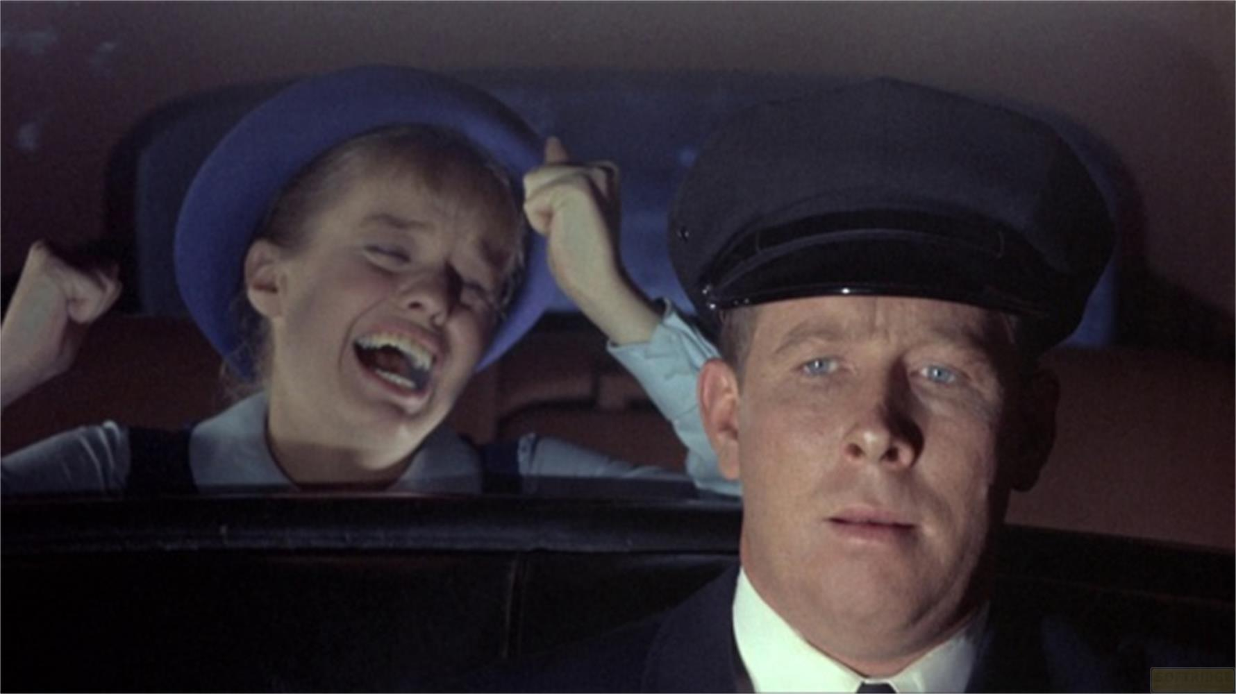 Un uomo con berretto da autista guarda avanti; una ragazza con cappello blu ride istericamente.